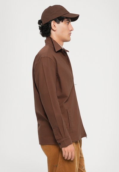 Un jeune homme portant une casquette marron, une veste zippée marron et un pantalon en velours côtelé clair se tenant de profil devant un fond blanc.