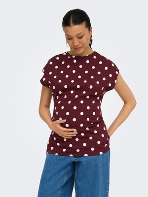 Pregnant woman gently cradling belly, wearing maroon polka dot top and blue jeans, with hair pulled back and hoop earrings.