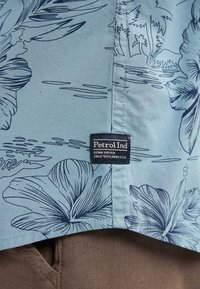 Camisa de manga corta azul claro con estampado floral en azul marino, costuras cosidas y una etiqueta negra "Petrol Ind" en el dobladillo.