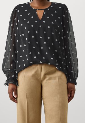 Blouse noire transparente à manches longues avec des pois blancs et des poignets froncés, associée à un pantalon beige taille haute sur une personne debout.