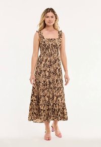 Maxi-jurk in een bruin-beige abstract patroon, met een gerimpeld lijfje, gerimpelde bandjes en een gelaagde rok. Gecombineerd met roze sandalen met hak.