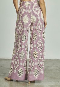 Pantaloni viola a zampa d'elefante con intricate ricami in pizzo bianco a motivo diamante e floreale, dotati di tasche e di una vita alta.
