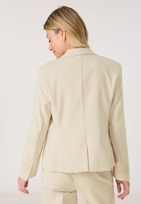 Hellbeige Blazer mit taillierter Passform, einem Rückenschlitz und drei Knöpfen an den Ärmeln. Glattes Material mit subtiler Textur und strukturiertem Design.