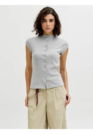 Kvinde med krøllet mørkt hår iført en lysegrå kortærmet knapbluse og beige vidde bukser med et rødt reb bælte.