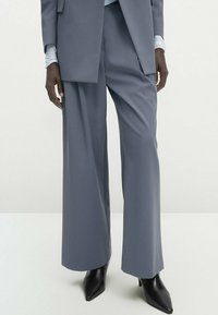 Grijze op maat gemaakte wijde pantalon van gladde stof, gecombineerd met zwarte puntige hakken. De outfit bevat een lichtblauw lang mouwtop eronder.