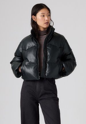 Femme aux cheveux foncés porte une doudoune noire sur un pull à col roulé rayé et un pantalon noir, regardant vers sa gauche avec les mains dans les poches de la veste.