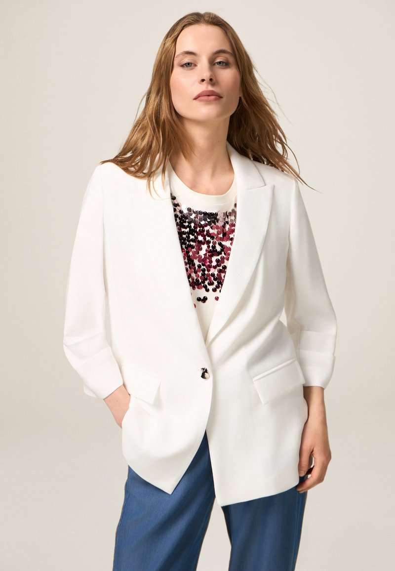 Blazer blanco con dos bolsillos frontales, cierre de botón simple, combinado con una blusa crema con acentos de lentejuelas negras y burdeos, usado con pantalones azules.
