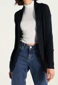 Schwarze, offen tragbare Strickjacke mit langen Ärmeln, aus glattem Material. Kombiniert mit einem strukturierten, eng anliegenden weißen Oberteil und hoch geschnittenen blauen Jeans.