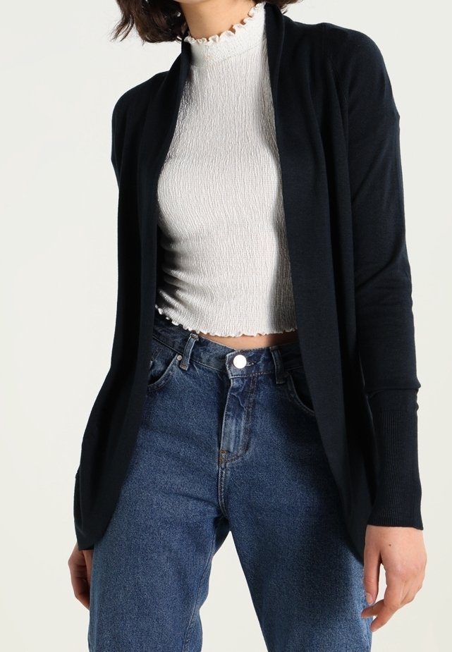 Schwarze, offen tragbare Strickjacke mit langen Ärmeln, aus glattem Material. Kombiniert mit einem strukturierten, eng anliegenden weißen Oberteil und hoch geschnittenen blauen Jeans.