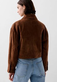 Kvindelig iført en cropped brun ruskindsjakke og lyseblå højtaljede jeans, vist fra ryggen mod en ensfarvet baggrund.