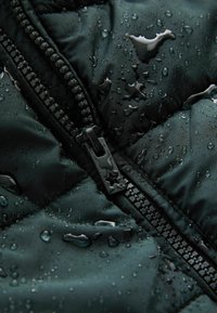 Vesikäiv dark green puffer-takki musta vetoketjulla. Pinnassa on vesipisaroita, mikä viittaa kosteudenkestävyyteen ja tekstuuriin.