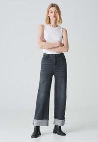 Jeans in denim scuro a gamba larga con risvolti e orli grigi a contrasto, abbinati a un top bianco senza maniche e stivaletti neri alla caviglia.