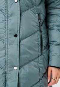 Gevoerde tealjack met een chevrondesign, voorzien van drukknoopjes en een ritszak aan de zijkant. Glad materiaal met glanzende accenten.