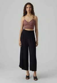 Mulher com cabelo cacheado vestindo um top cropped de renda mauve, calças largas pretas e sandálias de salto pretas, de pé contra um fundo cinza.