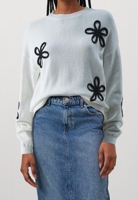 Person som bär en vit tröja med svarta blomapplikationer och en blå denimkjol, stående mot en enkel neutral bakgrund.