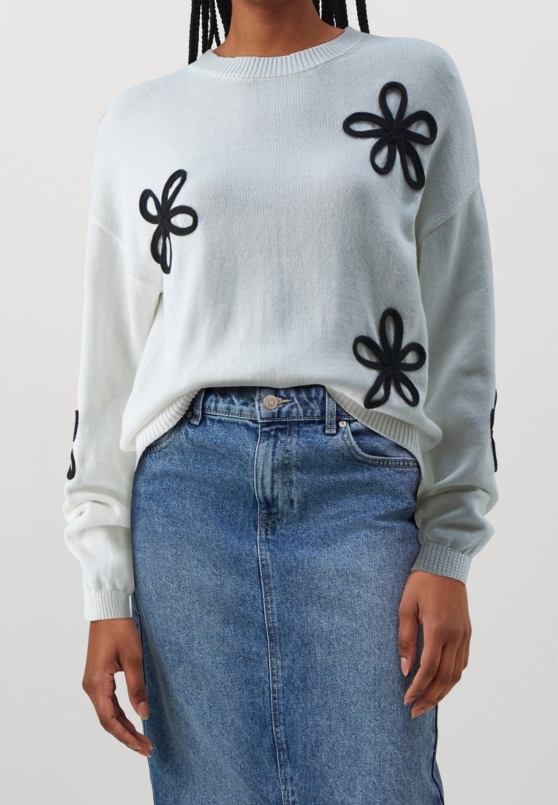 Person som bär en vit tröja med svarta blomapplikationer och en blå denimkjol, stående mot en enkel neutral bakgrund.