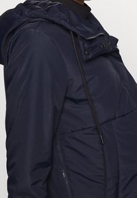 Marinblå puffjacka med strukturerad yta, justerbar huva, knappstängning fram och sidofickor med dragkedja för funktionalitet och värme.