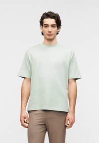 Ανοιχτό πράσινο βαμβακερό μπλουζάκι με στρογγυλή λαιμόκοψη και κοντά μανίκια, συνδυασμένο με καφέ παντελόνια. Απλός σχεδιασμός, άνετη εφαρμογή.