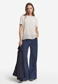 Vit kortärmad blus, mjukt tyg, avslappnad passform. Matchad med mörkblå vida jeans och en marinblå jacka, hålls i ena handen.