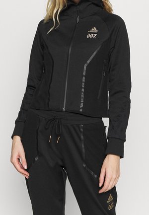 Kvinde iført sort Adidas 007 træningssæt med lynlåstrøje og snørede bukser, med gyldne Adidas- og 007-logoer.