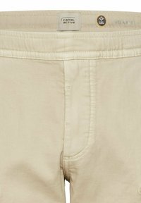 Beige Stretchhosen mit entspannter Passform, die ein sichtbares Taillenetikett, eine Fronttasche und genähte Details aufweisen.