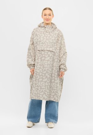 Donna sorridente, indossa una lunga giacca beige con cappuccio e motivo, con tasca frontale, jeans larghi blu e sneakers bianche, in piedi su sfondo bianco.