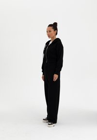 Musta vetoketjullinen huppari, jossa on tekstuuri, yhdistettynä löysiin mustiin collegehousuihin. Varustettu kiristysnyörillä, joustavalla vyötäröllä ja rentoilla mustilla tennareilla.