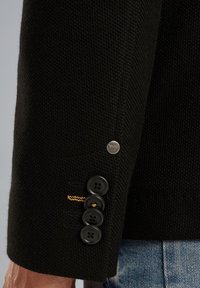 Zwarte blazer mouw met een textuurstof, drie zwarte knopen en een zilveren logo-accent bij de manchette met gele stiksels.