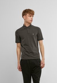Tmavošedé polo tričko s trojitej gombíkmi, krátkymi rukávmi a malým vyšitým logom na ľavej hrudi. Hladká bavlnená tkanina.