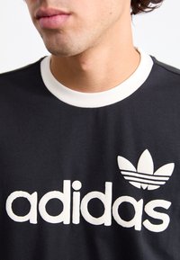 Camiseta negra de algodón, con un cuello redondo blanco, un prominente logotipo blanco de "adidas" y un emblema de tres hojas en el pecho.