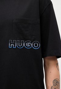 Tricou din bumbac negru, cu buzunar pe pieptul stâng și logo-ul brodat "HUGO" în degradé de albastru și alb. Mânici scurte și croială lejeră.