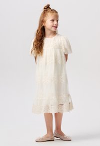 Witte kantjurk met korte pofmouwen, bloemenborduursel en een gelaagde zoom. Het kind draagt beige ballerina's met strikdetail.