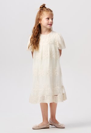 Weißes Spitzekleid mit kurzen Puffärmeln, floralem Stickerei und gestuftem Saum. Das Kind trägt beigen Ballettschuhe mit Schleifendetail.