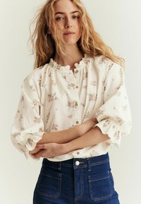 Ung kvinde med bølget blondt hår, der bærer hoop-øreringe, en hvid blomstrete knapbluse med flæseærmer og blå højtaljede jeans.