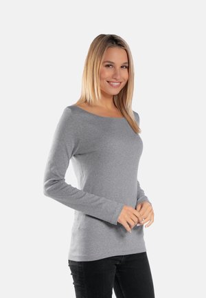 Uśmiechnięta kobieta o blond włosach, ubrana w dopasowaną szarą bluzkę z długim rękawem i czarne spodnie, stojąca na jasnym, jednolitym tle.