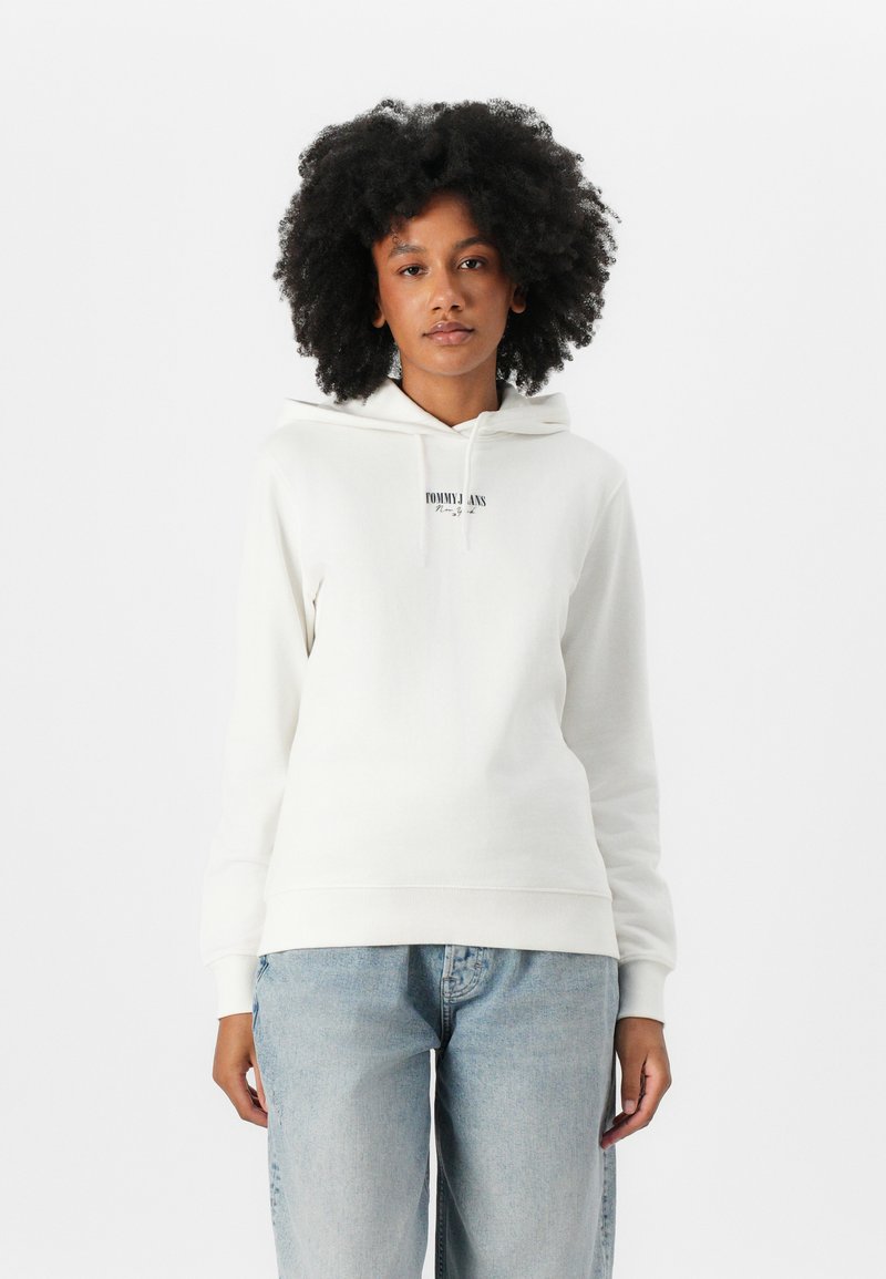 Witte hoodie van zacht materiaal met een kangoeroezak, een capuchon met trekkoord en een klein zwart logo op de borst. Gepaarde met lichtblauwe spijkerbroeken.