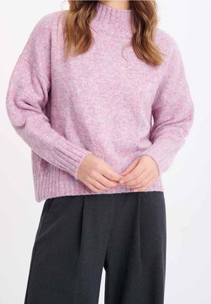 Vrouw draagt een lichtpaars gebreide trui met geribbelde mansetten en kraag, gecombineerd met losse zwarte broek, handen samengevouwen ter hoogte van de taille.