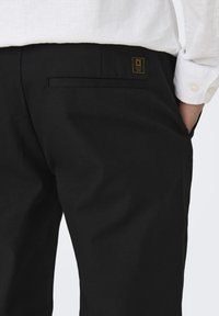 Pantaloni neri con una texture liscia, dotati di una tasca posteriore e una piccola etichetta con logo dorato. L'indumento è abbinato a una camicia bianca con bottoni.