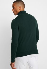 Pulover tricotat cu guler înalt de culoare verde închis, cu manșete și guler cu nervuri, având un design ajustat și o textură netedă, purtat peste pantaloni albi.