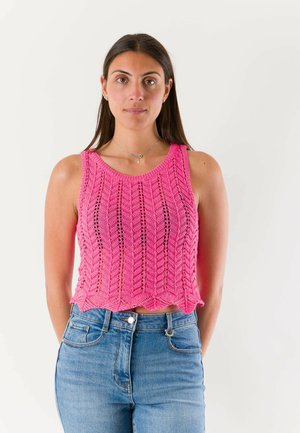 Donna con lunghi capelli scuri che indossa un top rosa lavorato a maglia senza maniche e jeans blu, in piedi su uno sfondo bianco uniforme.