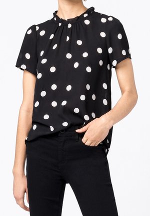 Sort kortærmet bluse med hvide polkaprikker og flæsekrave, båret ind i sorte bukser, hånd i lommen.