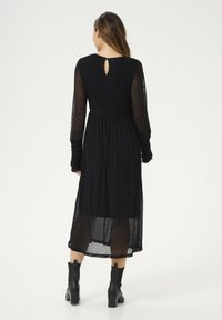 Svart långärmad klänning med smockat liv, transparenta ärmar och en detalj med nyckelhål i ryggen, med en flödande kjol och texturerad tyg.