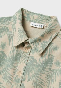 Camicia con colletto in tessuto beige con motivi di foglie tropicali verdi, due bottoni beige sul davanti e un'etichetta di cura all'interno del colletto.