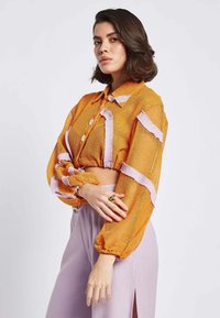 Giacca cropped arancione in rete con accenti rosa e maniche a sbuffo, dotata di colletto e bottoni frontali. Indossata con pantaloni lavanda a vita alta.