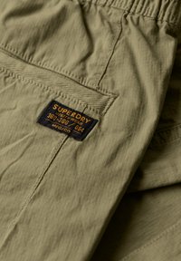 Olijfgroen lichtgewicht stof met een subtiele ribbelstructuur. Voorzien van een zwart label met gele tekst, genaaid in het taillegebied.