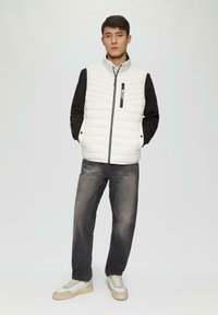 Weiße Pufferweste über einem schwarzen Langarmshirt, kombiniert mit grauen Jeans und beigen Sneakers. Bemerkenswerte Merkmale sind die gesteppten Texturen und Reißverschlussdetails.