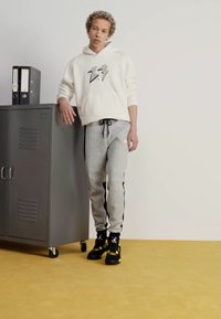 Witte hoodie met bliksemdesign, grijze sweatpants met zwarte zijkanten, en zwart-gele sneakers, geposeerd naast een grijze locker.