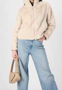 Beige, faux fur jacket with a high collar and zip closure. Paired with light blue wide-leg jeans and a small beige bag. Smooth texture.