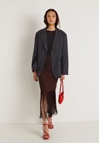 Grijze oversized blazer met knoopsluiting, gedragen over een bruine jurk met franjes, gecombineerd met rode accessoires en sandalen met bandjes.