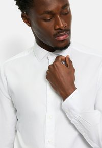 Weißes Hemd mit spitzem Kragen, langen Ärmeln und geknöpften Manschetten. Der Stoff wirkt glatt mit einer subtilen Textur.